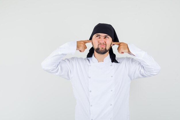 Chef macho tapando los oídos con los dedos en uniforme blanco y mirando aburrido. vista frontal.