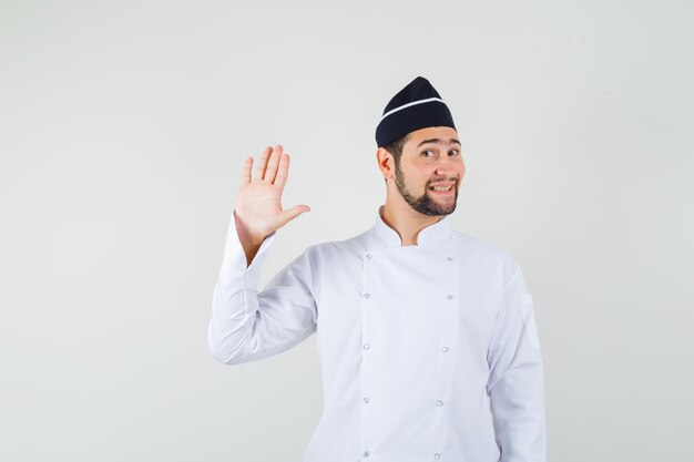 Chef masculino agitando la mano para saludar en uniforme blanco y mirando lindo, vista frontal.