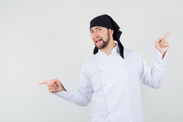 Chef masculino apuntando con el dedo hacia arriba y hacia abajo en uniforme blanco y mirando indeciso, vista frontal.