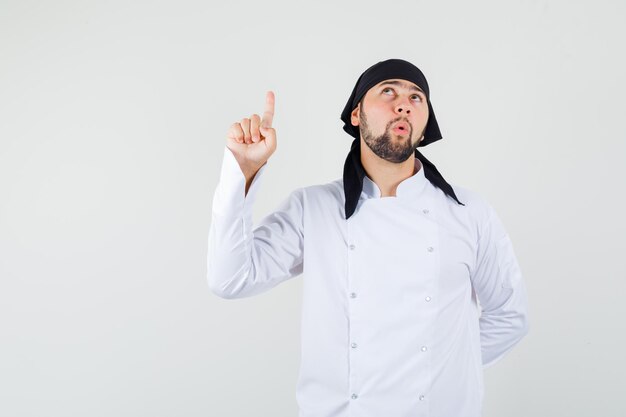 Chef masculino apuntando con el dedo hacia arriba en uniforme blanco y mirando enfocado, vista frontal.