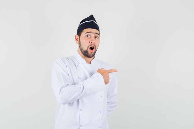 Chef masculino apuntando hacia un lado con uniforme blanco y mirando curioso, vista frontal.