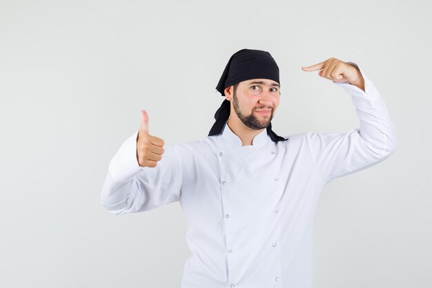 Chef masculino apuntando a su pañuelo con el pulgar hacia arriba en la vista frontal uniforme blanco.