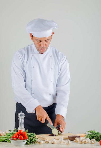 Chef masculino cortando berenjenas sobre tabla de madera en la cocina en uniforme, sombrero y delantal