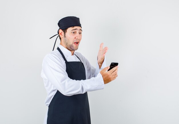 Chef masculino enojarse en videollamada en uniforme, delantal, vista frontal.