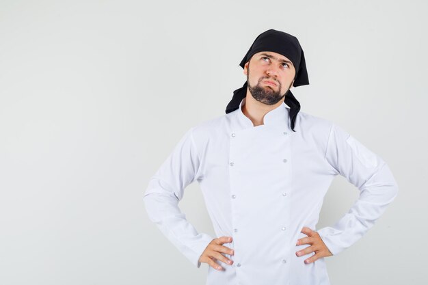Chef masculino mirando hacia arriba con las manos en la cintura en uniforme blanco y mirando indeciso, vista frontal.