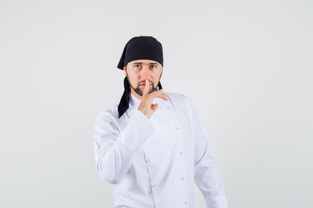 Chef masculino mostrando gesto de silencio en uniforme blanco y mirando con cuidado. vista frontal.