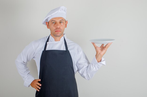 Chef masculino sosteniendo el plato y poniendo la mano en la cintura con sombrero, delantal y uniforme