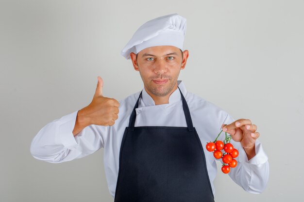 Chef masculino sosteniendo tomates y mostrando los pulgares para arriba con sombrero, delantal y uniforme