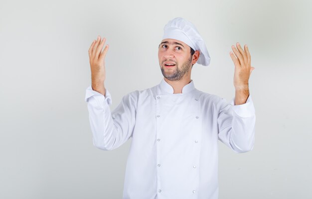 Chef masculino en uniforme blanco manteniendo las manos levantadas y mirando agradecido