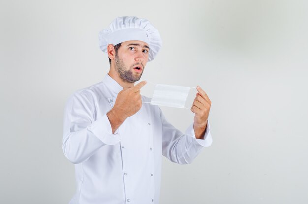 Chef masculino en uniforme blanco sosteniendo una máscara médica y mirando vacilante