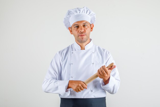 Chef masculino en uniforme, delantal y sombrero con rodillo y mirando confiado