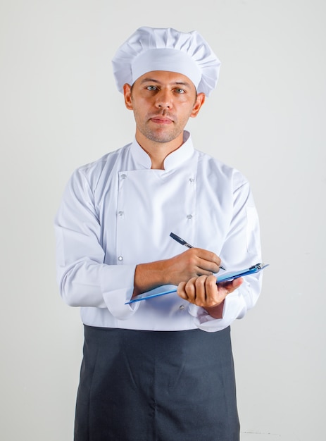 Chef masculino en uniforme, delantal y sombrero tomando notas en el portapapeles