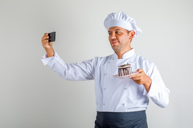 Chef masculino en uniforme, delantal y sombrero tomando selfie con pastel de postre