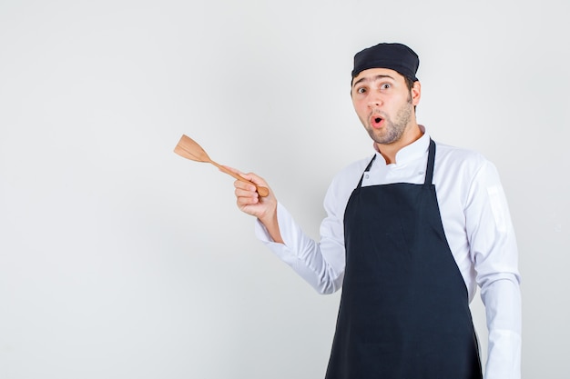 Chef masculino en uniforme, delantal sosteniendo una espátula de madera y mirando sorprendido, vista frontal.