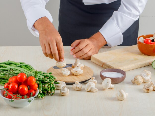 Chef masculino en uniforme y delantal tomando hongos para picar en la cocina