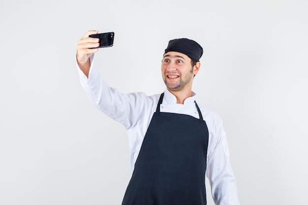 Chef masculino en uniforme, delantal tomando selfie en smartphone y mirando alegre, vista frontal.