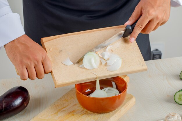 Chef masculino en uniforme y delantal vertiendo cebollas picadas en un tazón en la cocina