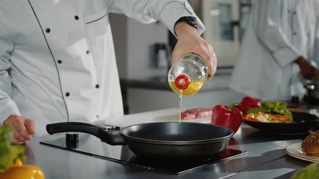 Chef masculino vertiendo aceite de girasol de una botella en una sartén en la estufa, preparando líquido para freír para hacer una deliciosa comida gourmet. Hombre profesional cocinando recetas de carne y vegetales orgánicos. De cerca.