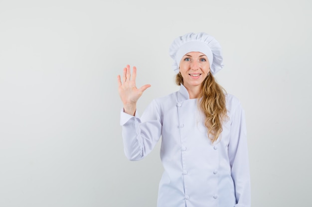 Chef mujer agitando la mano para decir hola o adiós en uniforme blanco y mirando alegre