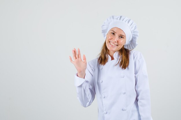 Chef mujer agitando la mano para saludar en uniforme blanco y mirando contento. vista frontal.