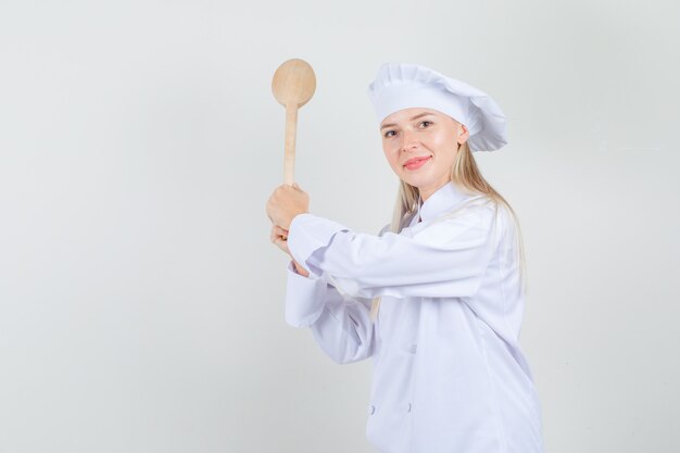 Chef mujer amenazando con cuchara de madera en uniforme blanco y mirando alegre