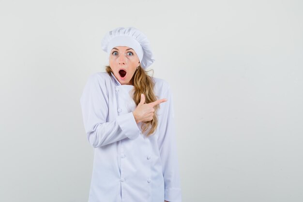 Chef mujer apuntando hacia afuera en uniforme blanco y mirando consternado