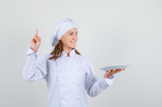 Chef mujer apuntando hacia arriba mientras sostiene el plato en uniforme blanco y mirando contento