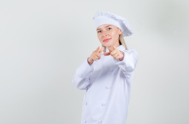 Chef mujer apuntando a la cámara con gesto de pistola en uniforme blanco y mirando alegre