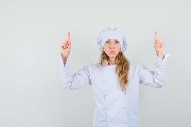 Chef mujer apuntando con el dedo hacia arriba y haciendo pucheros labios en uniforme blanco