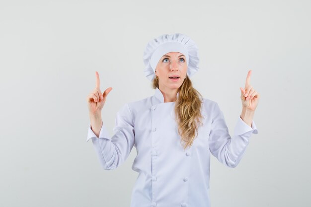 Chef mujer apuntando con el dedo hacia arriba en uniforme blanco y luciendo optimista.