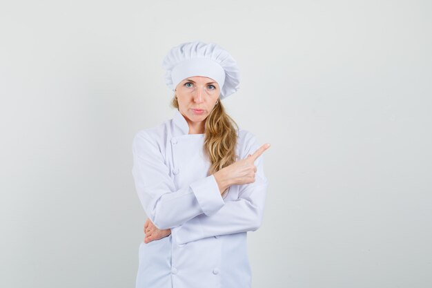 Chef mujer apuntando hacia fuera en uniforme blanco y mirando dudoso