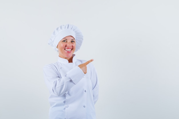 Chef mujer apuntando a un lado en uniforme blanco y mirando alegre