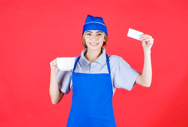 Chef mujer en delantal azul sosteniendo una taza de fideos de cerámica blanca y presentando su tarjeta de visita