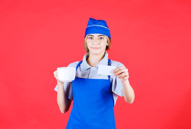 Chef mujer en delantal azul sosteniendo una taza de fideos de cerámica blanca y presentando su tarjeta de visita