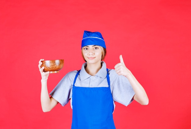 Chef mujer en delantal azul sosteniendo una taza de fideos de cobre chino y mostrando signo de mano de disfrute