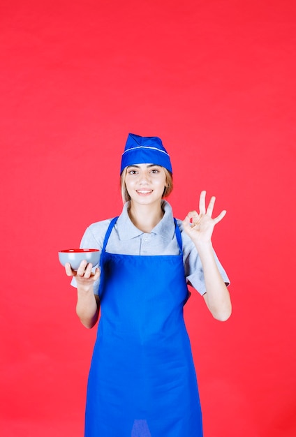 Chef mujer en delantal azul sosteniendo una taza de fideos y mostrando signo de mano de satisfacción