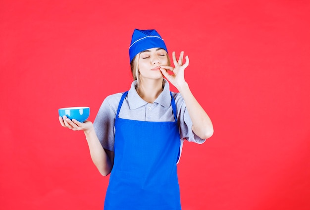 Chef mujer en delantal azul sosteniendo una taza de fideos y mostrando signo de mano de satisfacción