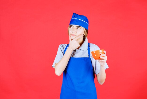 Chef mujer en delantal azul sosteniendo una taza de fideos y parece confundida y pensando en cómo hacerlo más delicioso