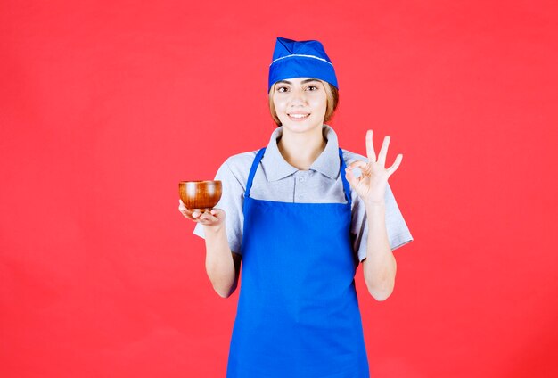 Chef mujer en delantal azul sosteniendo una taza de té de cobre chino y sintiéndose positivo sobre el sabor