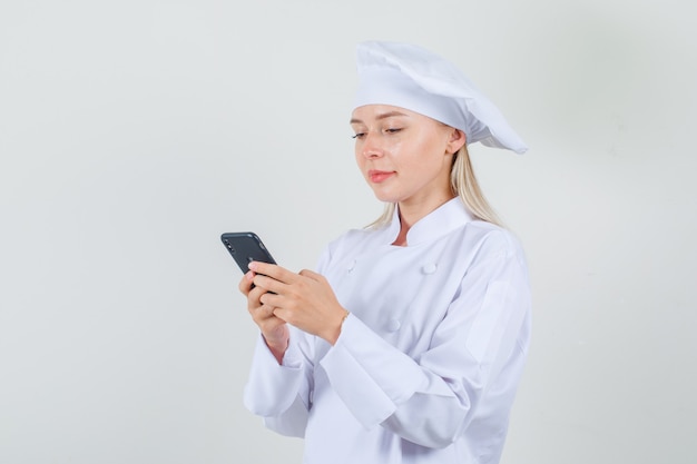 Chef mujer escribiendo en el teléfono inteligente y sonriendo en uniforme blanco