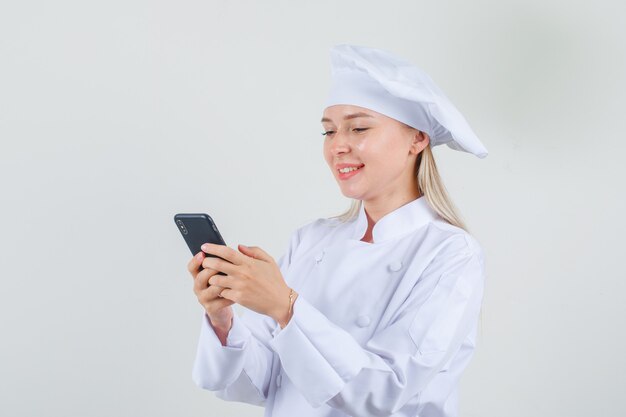 Chef mujer escribiendo en el teléfono inteligente y sonriendo en uniforme blanco