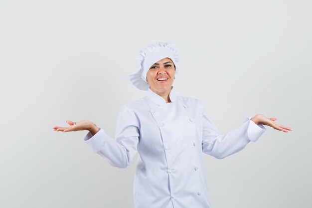 Chef mujer extendiendo las palmas a un lado en uniforme blanco y mirando contento.