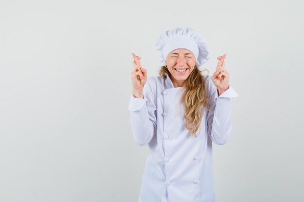 Chef mujer manteniendo los dedos cruzados en uniforme blanco y luciendo dichosa.