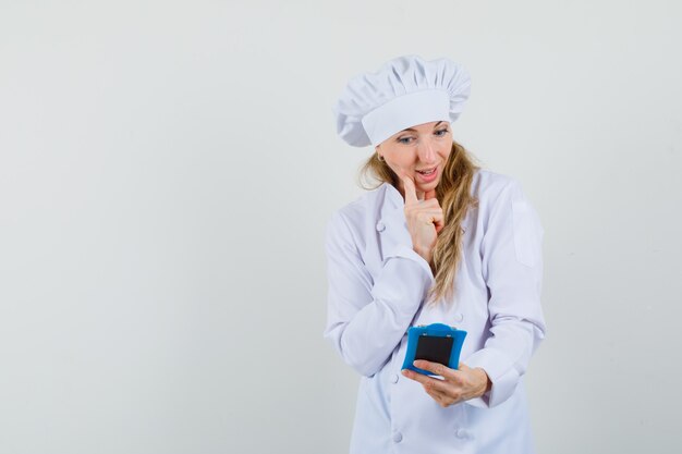 Chef mujer mirando notas en el portapapeles en uniforme blanco y mirando alegre