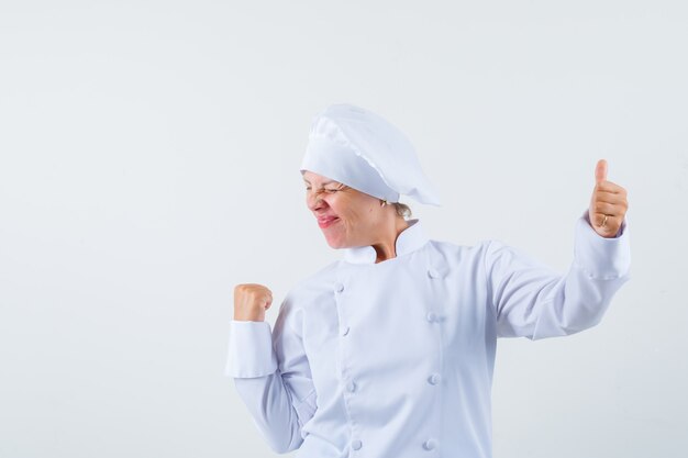 Chef mujer mostrando el pulgar hacia arriba mientras muestra el gesto ganador en uniforme blanco y mirando alegre