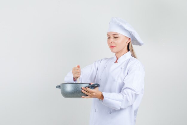 Chef mujer revolviendo la comida en una olla en uniforme blanco y mirando alegre. vista frontal.