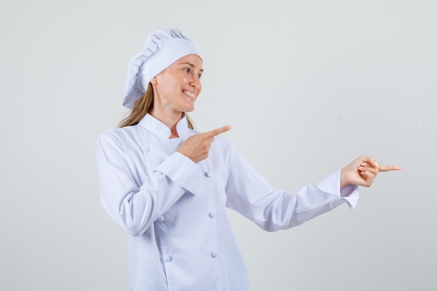 Chef mujer señalando con el dedo a los lados en uniforme blanco y mirando alegre. vista frontal.