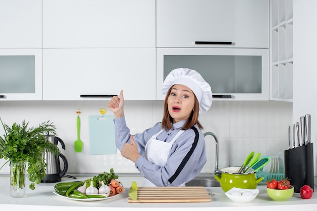 Chef mujer sorprendida y verduras frescas con equipo de cocina y haciendo un gesto de ok en el lado derecho en la cocina blanca