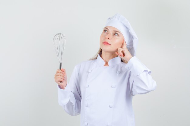 Chef mujer sosteniendo batidor y mirando soñadora en uniforme blanco