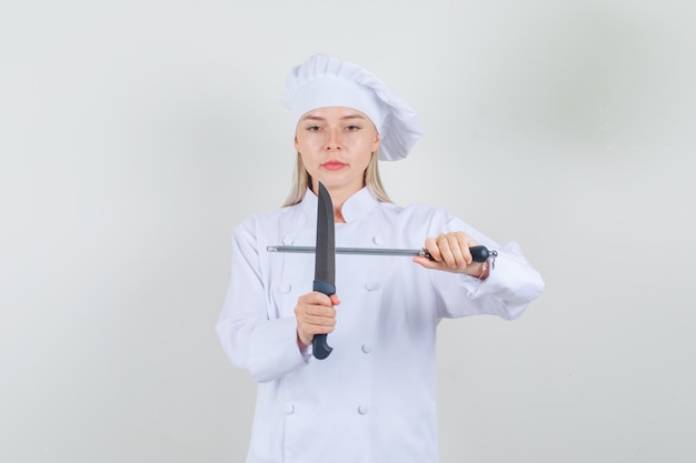 Chef mujer sosteniendo un cuchillo y un afilador en uniforme blanco y mirando serio